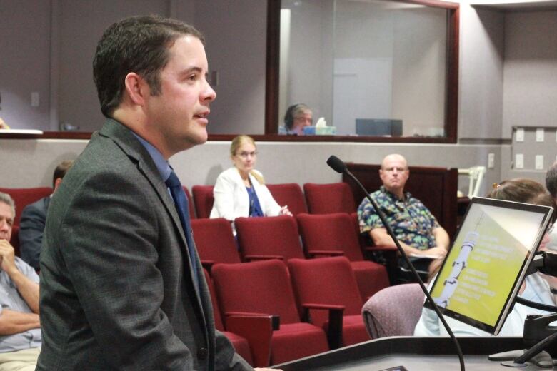 A profile shot man with short brown hair speaking at a podium with several people watching in red gallery seats in the background.