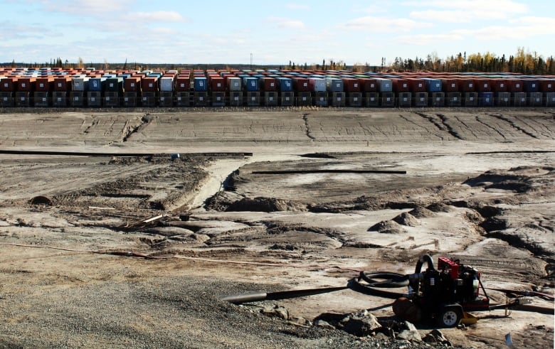 Rows of shipping containers sit on top of each other in the distance on a landscape that is sand and dirt. There is some machine that sits in the front.