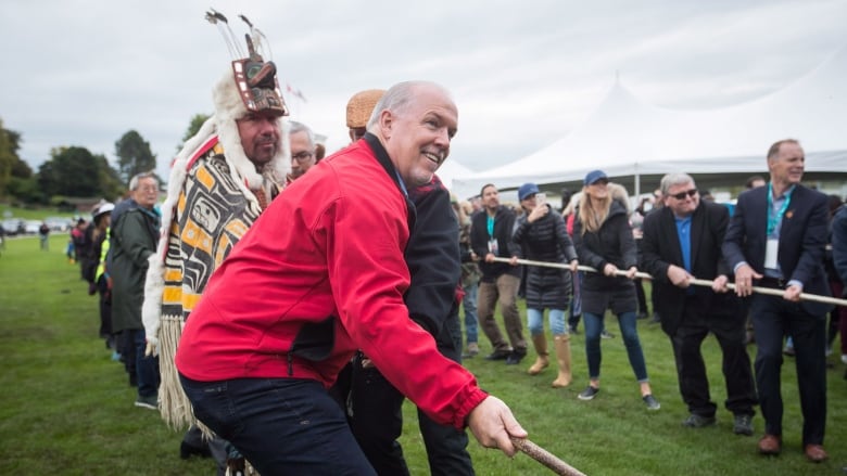 People, including a man in a red jacket next to a man in full Indigenous regalia, pull on poles to raise a totem 