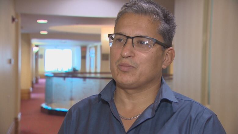 An Indigenous man with short cut grey hair is seen wearing half rim glassed and a blue button up 