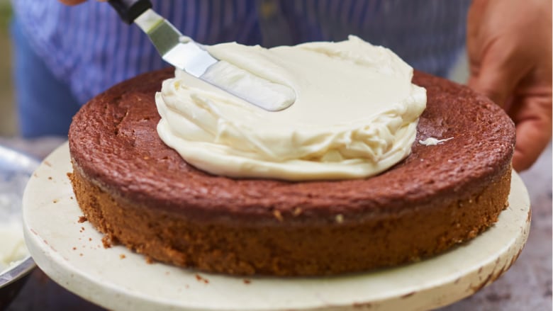 Closeup on a single-layer cake sitting on a white cake stand. A hand holds the side of the stand and a spatula spreads white icing on top of the cake. 