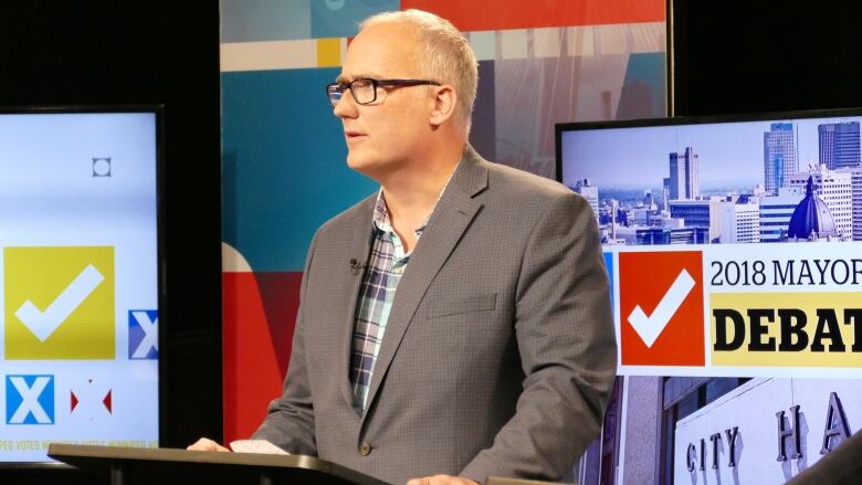 A man in a suit stands at a podium, in front of a TV screen with a graphic reading 