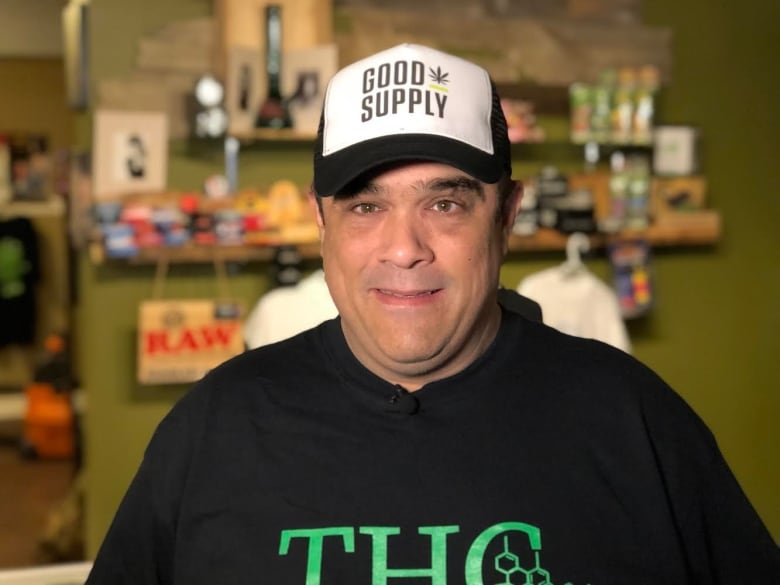 A man wearing a black hat and black t shirt standing inside a cannabis supply store. 