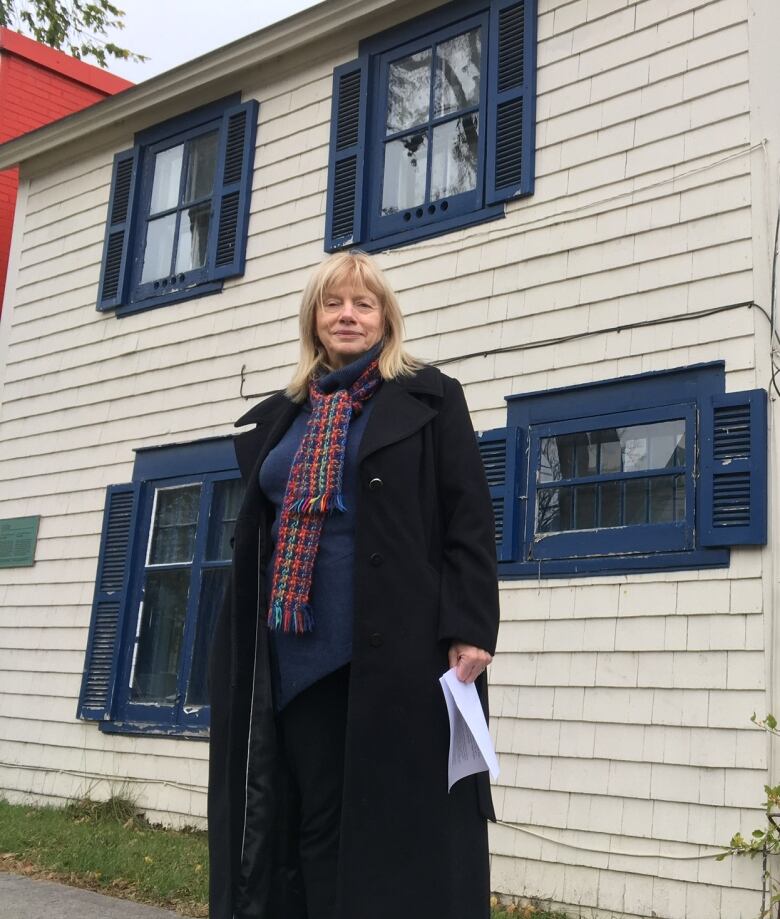 A woman in a long dark coat stands in front of a house.