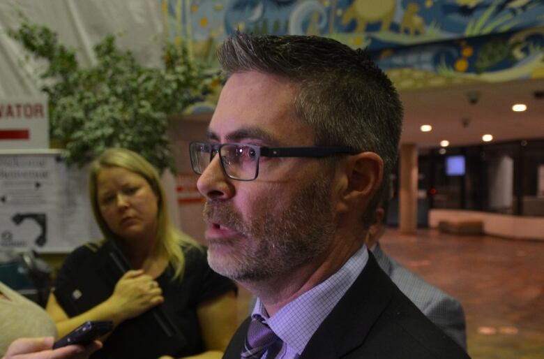 Close up photo of a man speaking into several microphones, with a reporter listening in the background. 