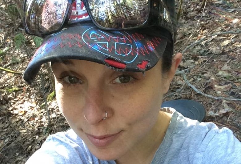 Photo of Ashley Simpson looking at the camera and smiling. She is wearing a ball cap on her head with sunglasses on the top of the rim. 