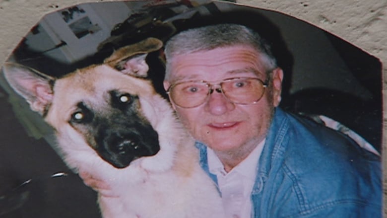 A man wearing glasses, a white shirt with a denim jacket hugs a German Shephard dog.