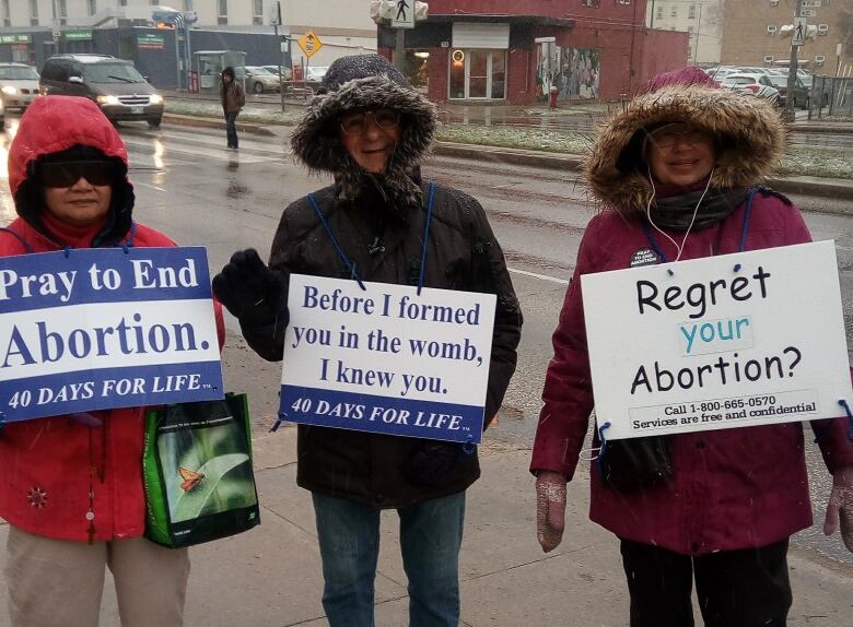 Three people stand along the side of the road, bundled in jackets, wearing signs that oppose abortions. 