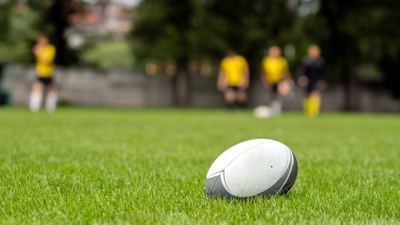 A rugby ball sits on the grass. Players are blurred in the background.