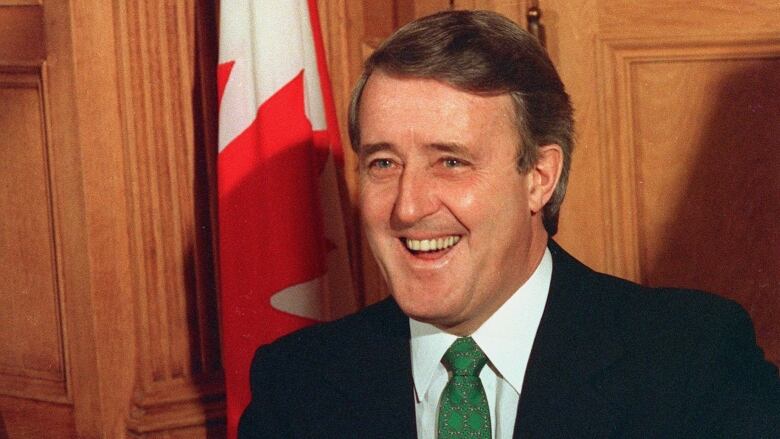 A man smiles with a Canadian flag hanging in the background.