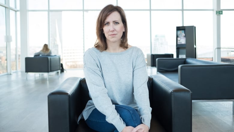 A woman sits in a black chair in a large room that has windows in the background.