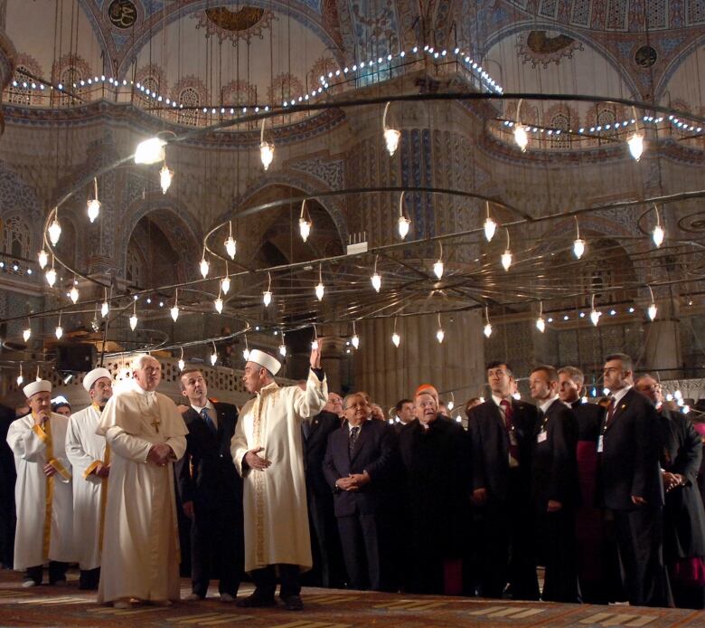 A man dressed in white robes walks through a mosque, with a group of men standing behind him.