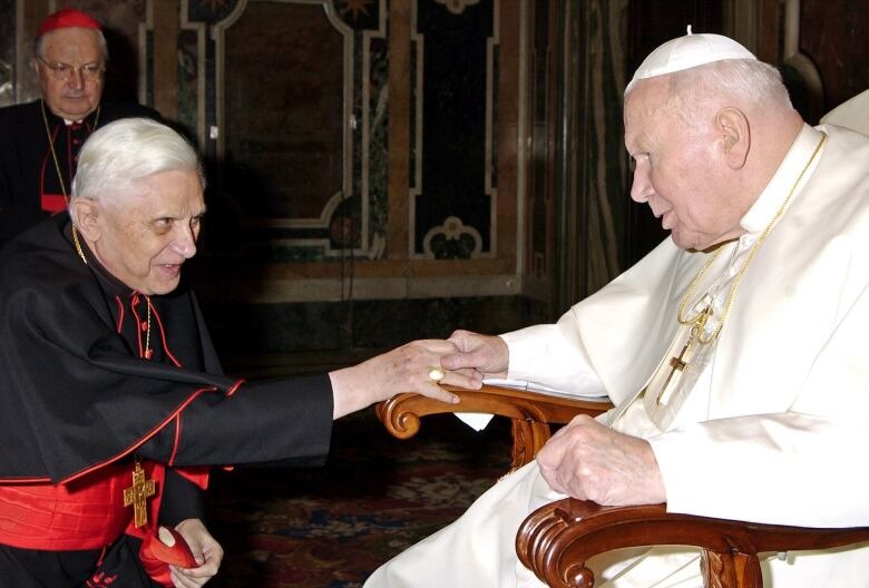 A man dressed in black and red robes shakes hands with a man dressed in white robes.