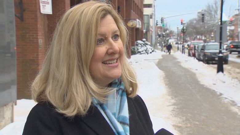 A woman in a black winter coat and blue scarf is looking off camera at someone interviewing her on a snowy sidewalk.