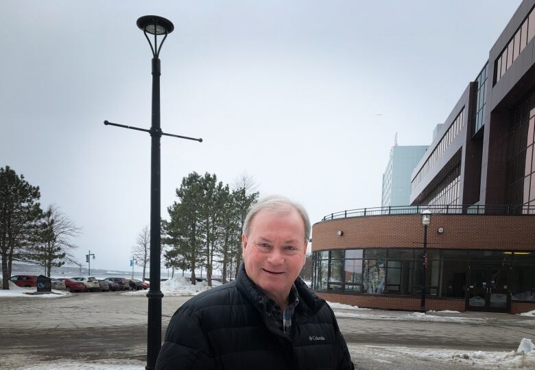 A man wearing a dark jacket stands in front of a building with some snow on the ground.