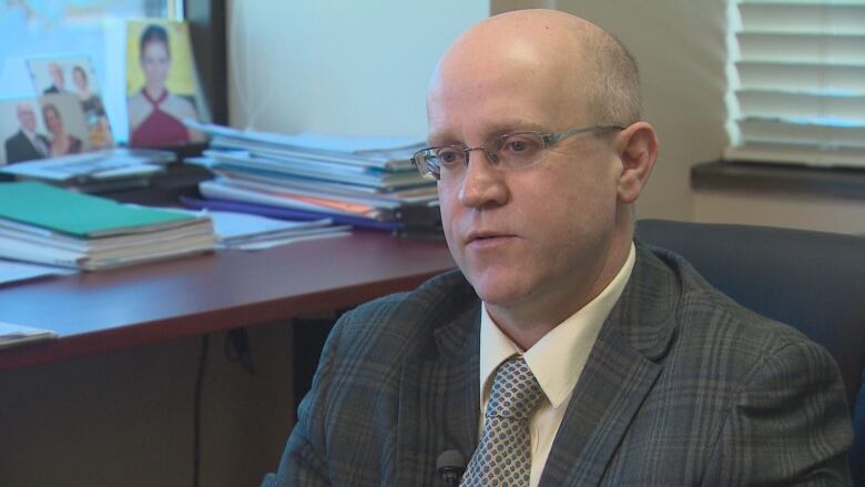 A bald man with glasses wearing a grey plaid suit is sitting in an office.