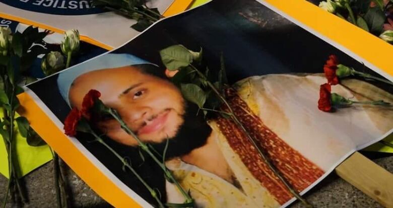 A picture of a man smiling at the camera, pasted on paper on top of a stick. Flowers are seen on top of the photo.Soleiman Faqiri was born on New Year's Day in Kabul, Afghanistan, in 1986 and came to Canada in 1993. According to his family, he was a straight-A student, captain of his high school rugby team and had a close and loving relationship with his four siblings and parents. 