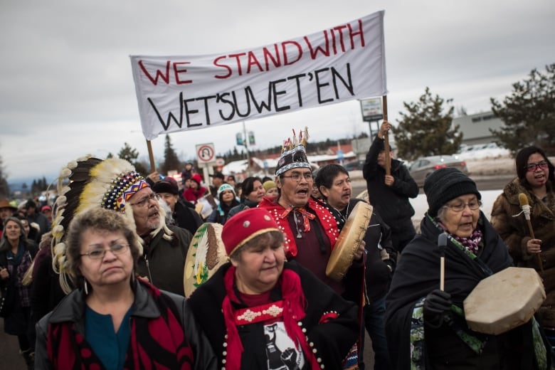 A group of people, some in First Nation's regalia and carrying drums, march beneath a sign saying 'We Stand with Wet'suwet'en.'