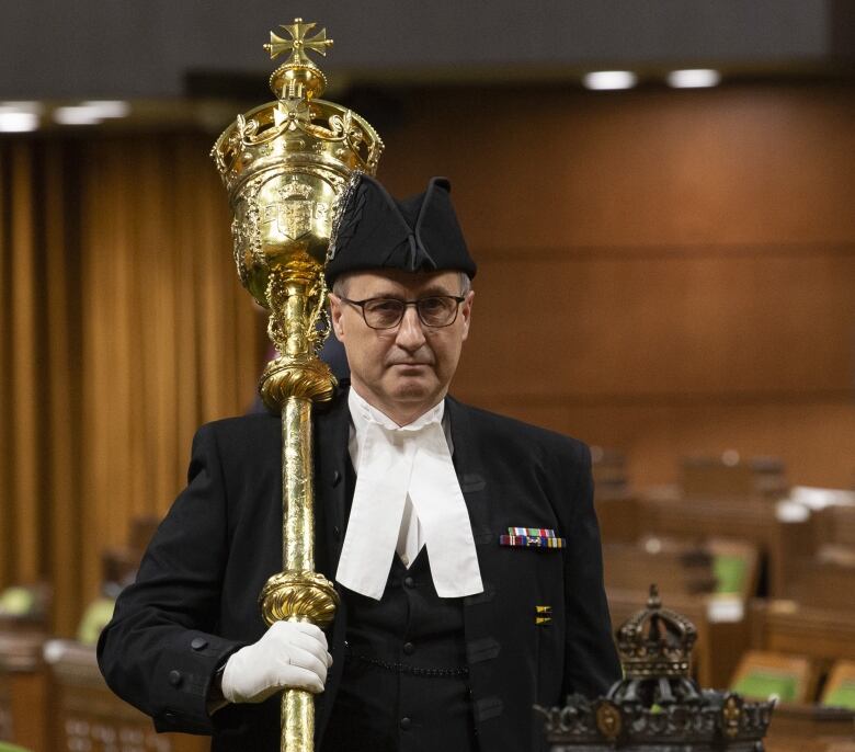 A man wearing a black suit and a traditional House of Commons white collar carries a gold ceremonial mace into the House.