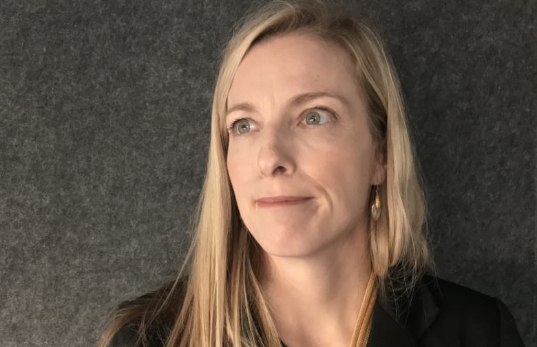 A woman poses in front of a grey wall, looking to the left of the camera.