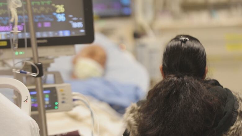 The back of a woman's head is shown in the foreground, in front of a blurry person in a hospital bed