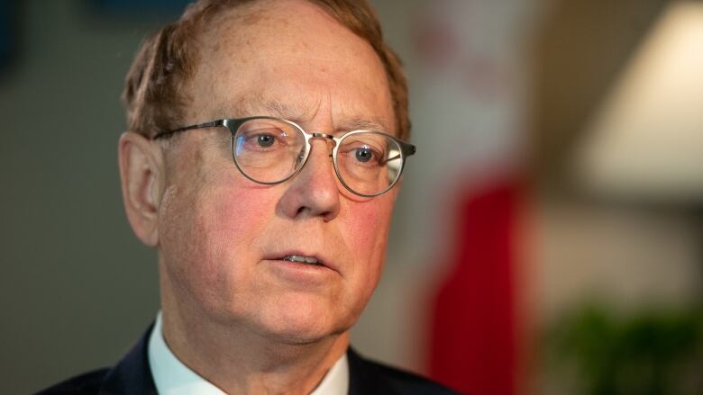 A close-up shot of a man in glasses. He's wearing a dark blazer, a white shirt, and a colorful red tie.