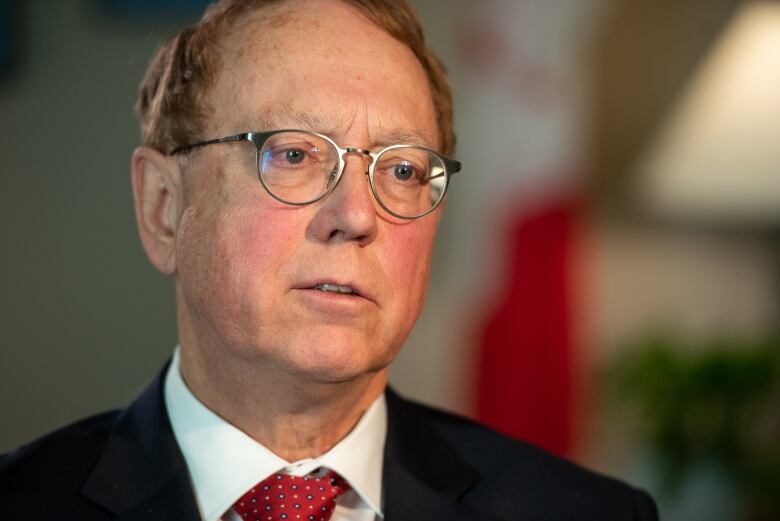 A close-up shot of a man in glasses. He's wearing a dark blazer, a white shirt, and a colorful red tie.