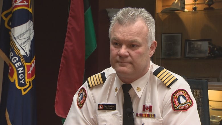 A white man with short grey hair is wearing a white uniform shirt with red Halifax Fire badges on his shoulders and black shoulder plates. There are flags behind him in shadows