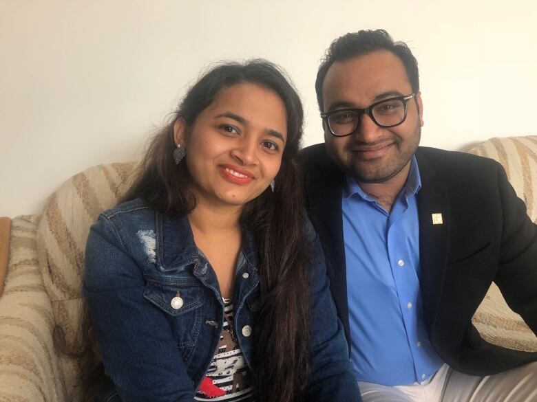 A married couple sits on their couch while smiling at the camera. The man's arm is around the woman.