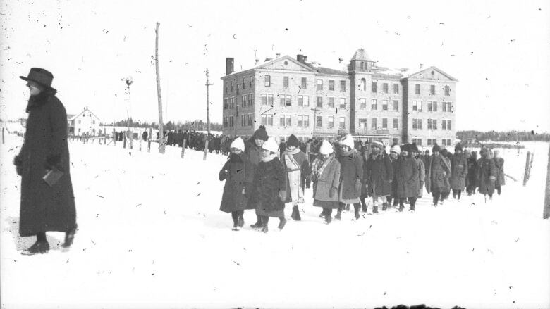 A black and white photo shows children in coats and hats walking through the snow in front of large brick building, following a person wearing a dark coat and wide-brim hat.