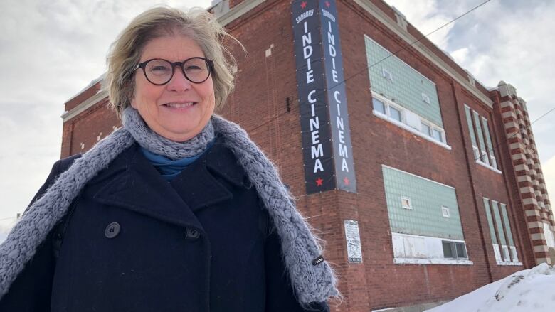 A smiling woman wearing a grey scarf and blue jacket stands in front of a brick building with an Indie Cinema sign