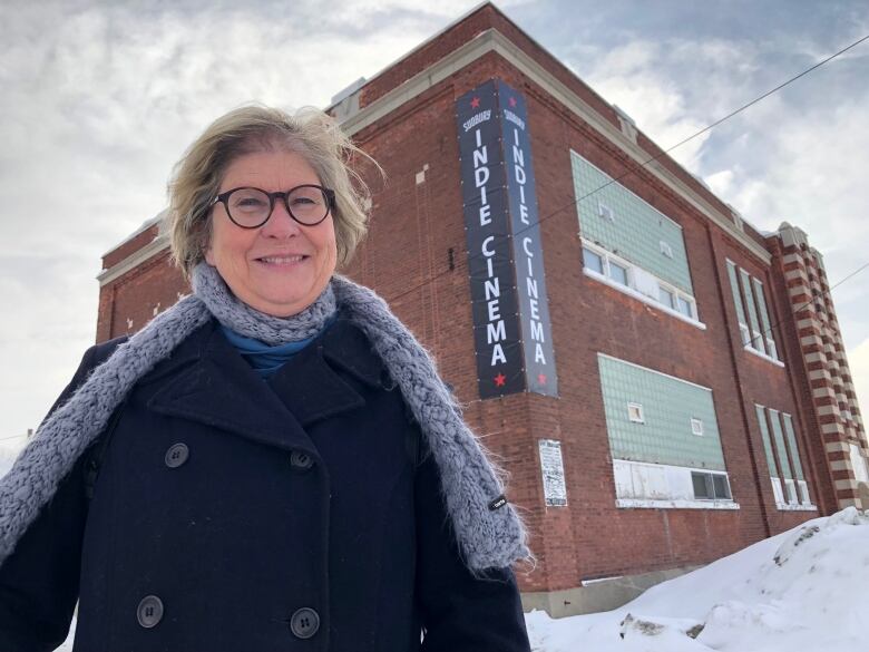 A smiling woman wearing a grey scarf and blue jacket stands in front of a brick building with an Indie Cinema sign