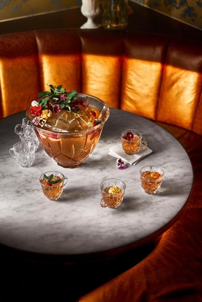 A crystal punch bowl filled with a golden orange punch. Small glasses of punch sit next to it. They're all on a marble table. 