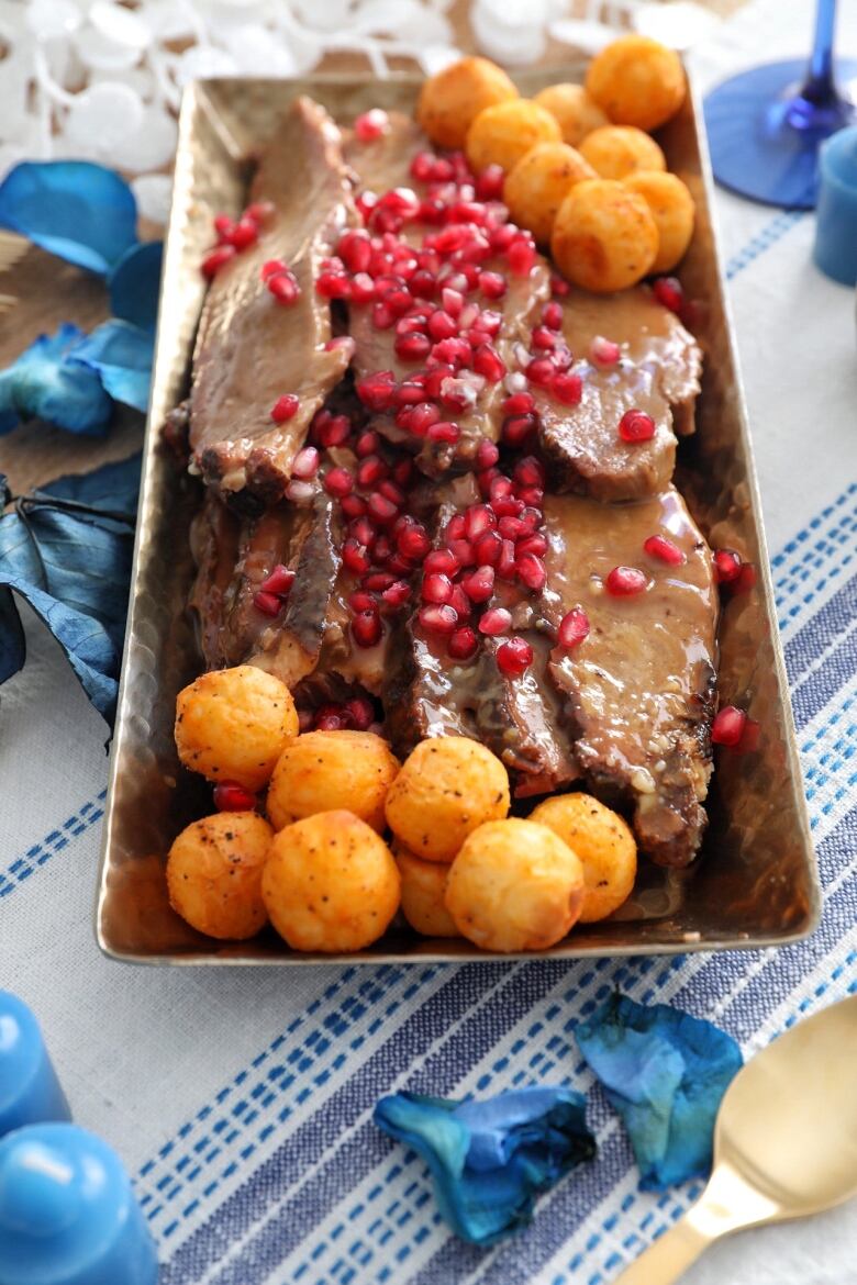 Closeup on a rectangular serving dish with brisket and mini potatoes with pomegranate arils on top. The decor on the table is blue and white.