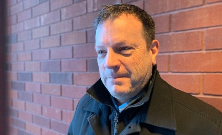 A man with short brown hair wearing a black jacket stands in front of a brick wall.