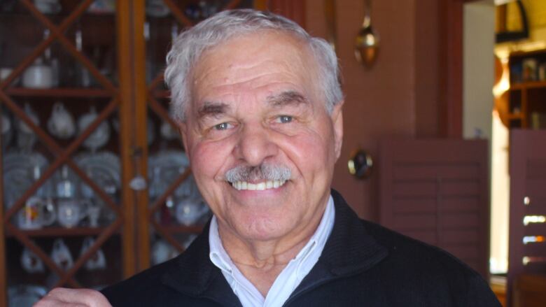 A man with short grey hair and a grey moustache smiles at the camera while holding up a photo of a bronze-coloured statute in front of a brick building. He is wearing a black sweater over a light coloured collared shirt and standing indoors.