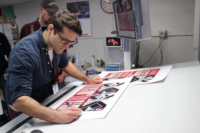 A man in glasses and a navy button-down shirt leans over publishing proofs on a drafting table, to scribble notes in the margin.  