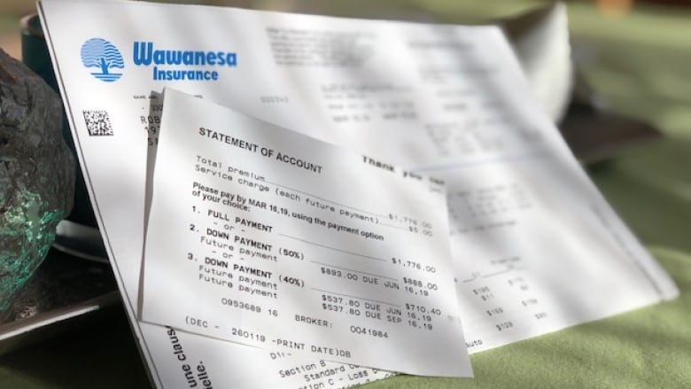 An invoice from Wawanesa Insurance is displayed on a cloth background. A small paper saying Statement of Account obscures personal details of the client. 