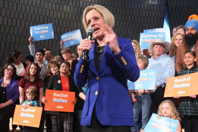 A politician speaks into a microphone, with sign-waving people behind her. 