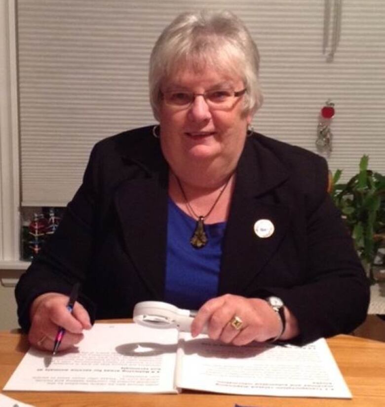 A woman with white hair, glasses and a dark suit jacket sits at a table and uses a magnifying glass to read.