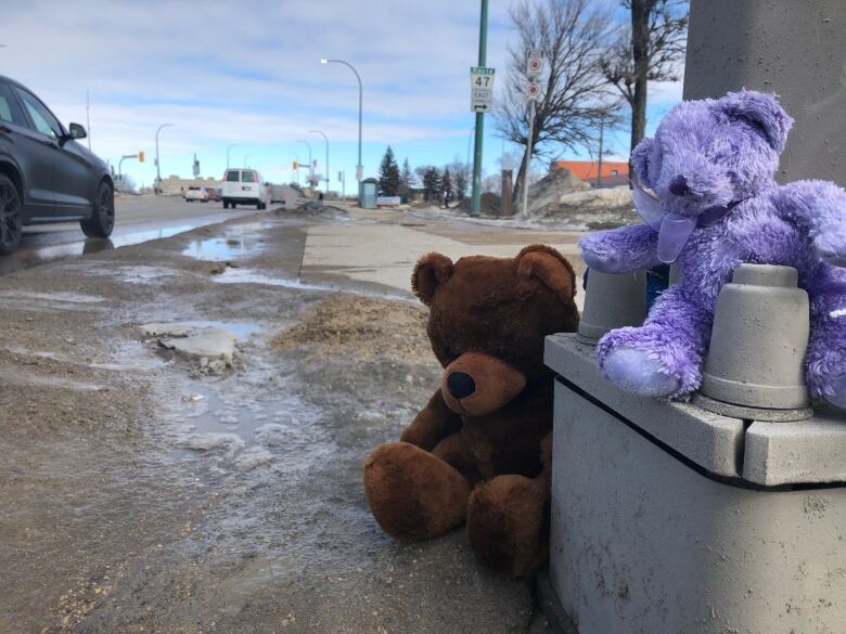 Two teddy bears, one brown and one purple, have been placed at the base of a light pole next to a busy street.