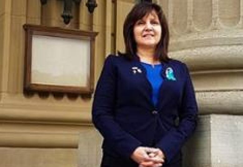 A dark-haired woman in a blue suit stands for photo beside a large vertical column outside the Alberta Legislature. 