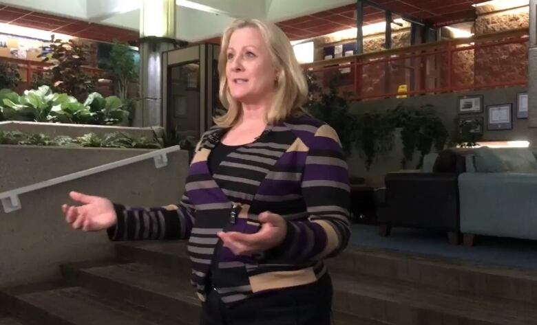 A blonde woman wearing a striped sweater stands inside a building, speaking to someone out of view.