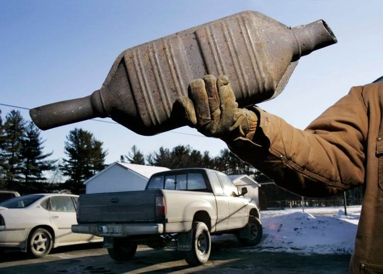 An unseen man holds up a catalytic converter, which is thick in the middle with pipes coming out either end.