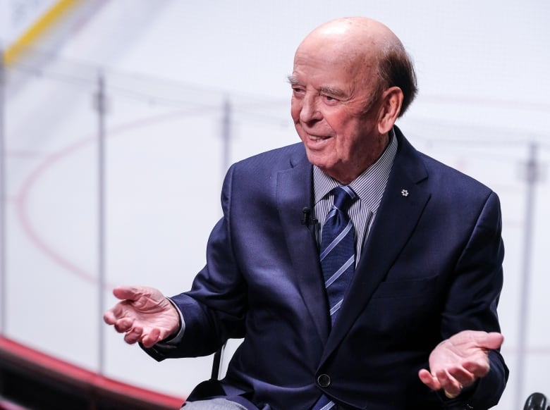 A man wearing a suit and tie smiles and raises both hands at his sides. Behind him is the surface of a hockey arena. 