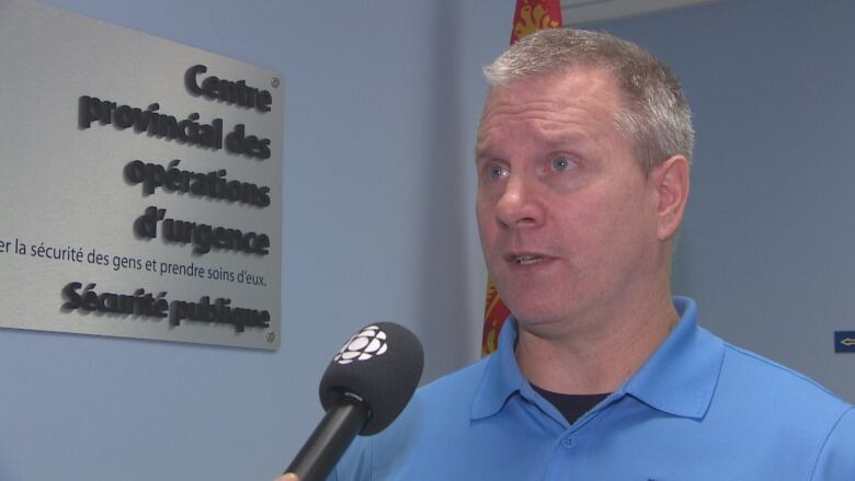 A man with short silver hair and a blue shirt speaks into a microphone with a CBC mic flash on it.