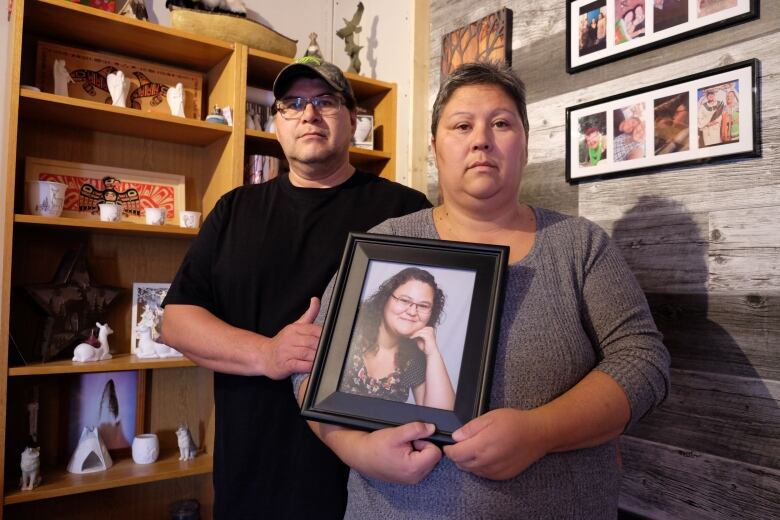 A man and a woman are pictured standing in front of a shelf, holding a framed photo of their daughter.