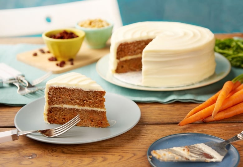 A carrot cake with one slice cut from it. It's sitting on a white plate on a wooden table. The slice of cake is on a blue plate in front of the full cake. 