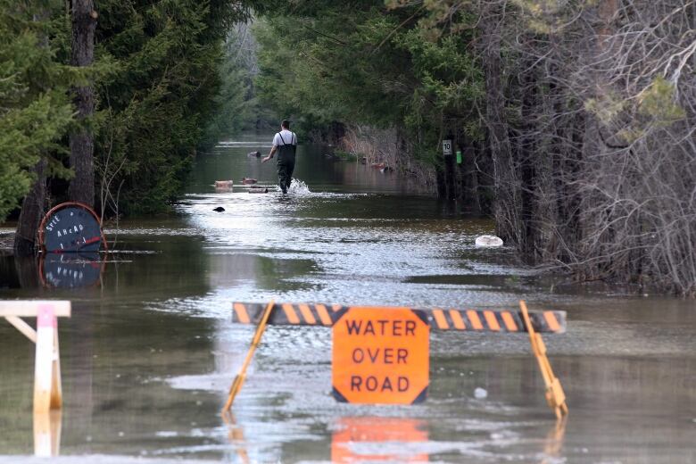 A man walks on a flooded road with a caution sign in the foreground of the image saying 