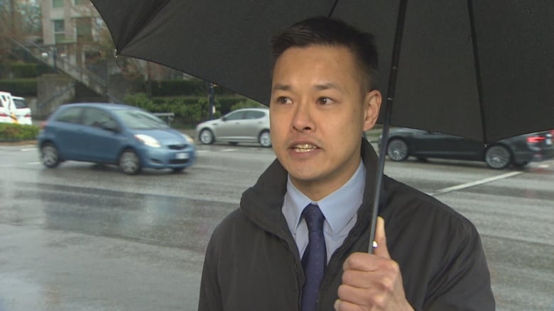 An East Asian man speaks underneath an umbrella.